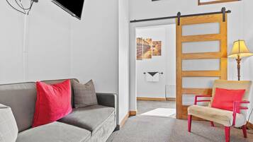 Uma sala de estar moderna com um sofá cinzento, almofadas vermelhas e cinzentas, uma poltrona vermelha e bege, uma porta de correr de madeira e um abajur.
