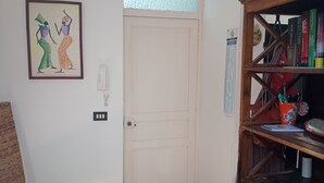Una habitación con una puerta blanca, una silla de mimbre, una estantería y un cuadro en la pared.