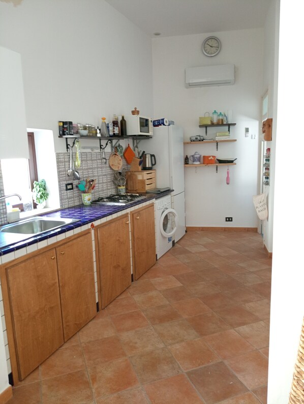 A kitchen with wooden cabinets, a sink, a microwave, and a washing machine. There are shelves with various kitchen items and a clock on the wall.
