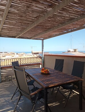 A covered outdoor seating area with a table and chairs, overlooking a cityscape and the sea.