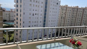 Ein Balkon mit einem Glastisch und einem blauen Kissen, mit Blick auf ein Wohngebiet mit weißen Mehrfamilienhäusern.