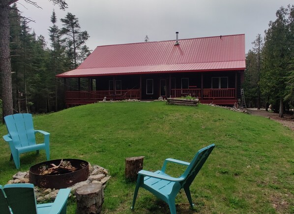 A cabin with a red roof, surrounded by a grassy area with two blue Adirondack chairs and a fire pit.
