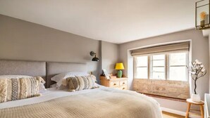 Ein Schlafzimmer mit einem großen Bett, einem Nachttisch mit Lampe, einem Fenster mit Vorhängen und einem kleinen Tisch mit Pflanze.