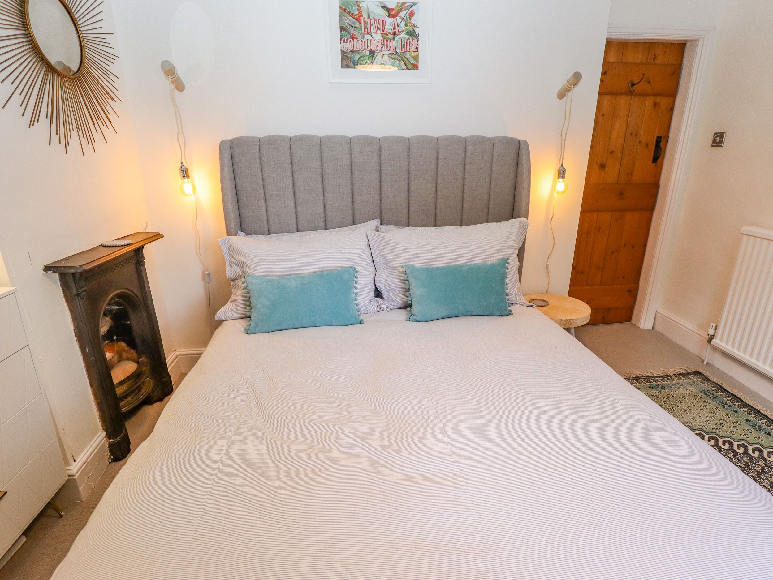 A bedroom with a large bed, two pillows, a headboard, a wall-mounted mirror, a framed picture, and a wooden door.