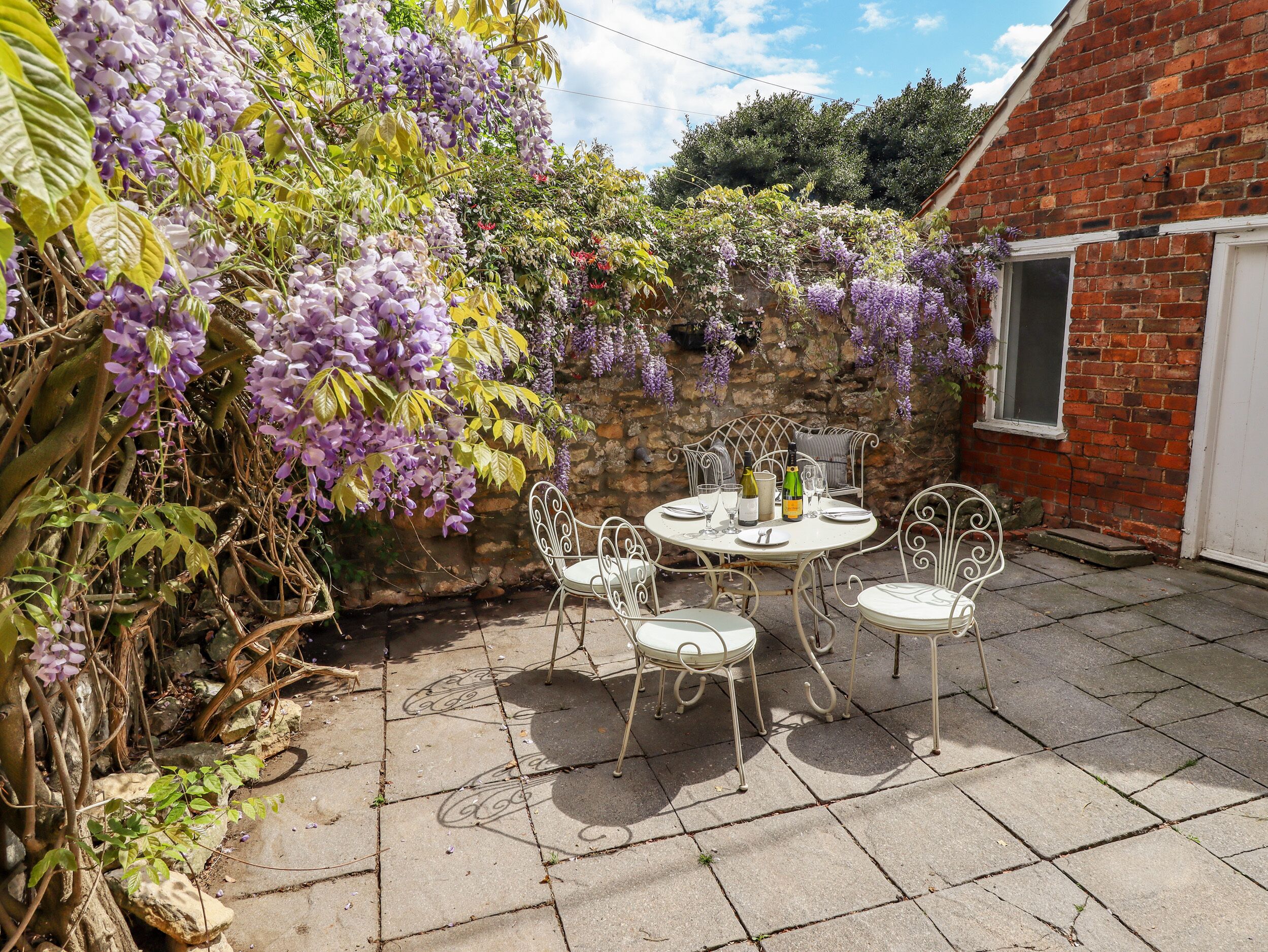 A patio with a round table and four chairs, surrounded by a brick wall and covered with purple wisteria.
