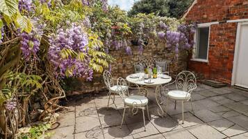 A patio with a round table and four chairs, surrounded by a brick wall and covered with purple wisteria.