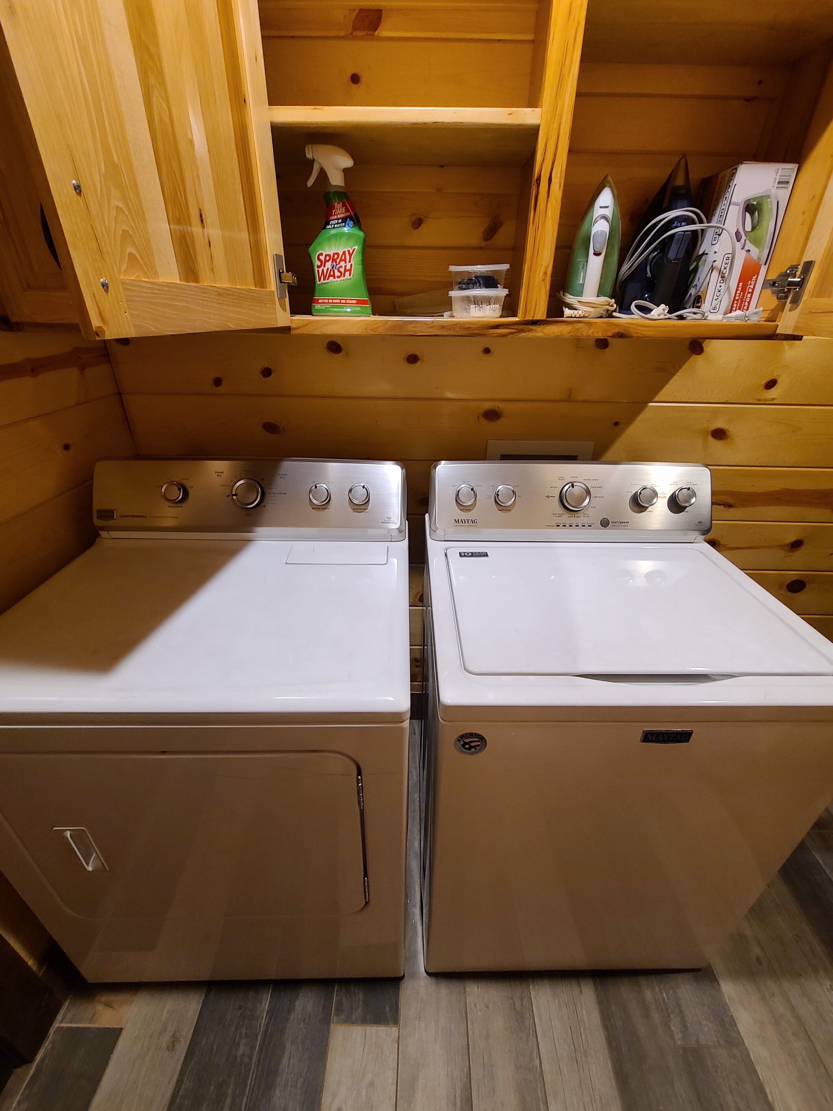 A laundry room with two white front-loading washers and dryers, wooden cabinets above, and a shelf with cleaning supplies.