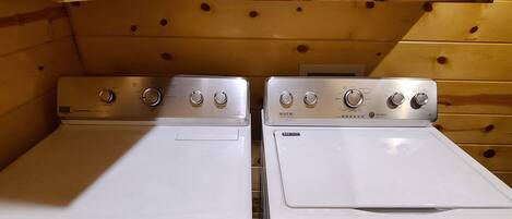 A laundry room with two white front-loading washers and dryers, wooden cabinets above, and a shelf with cleaning supplies.