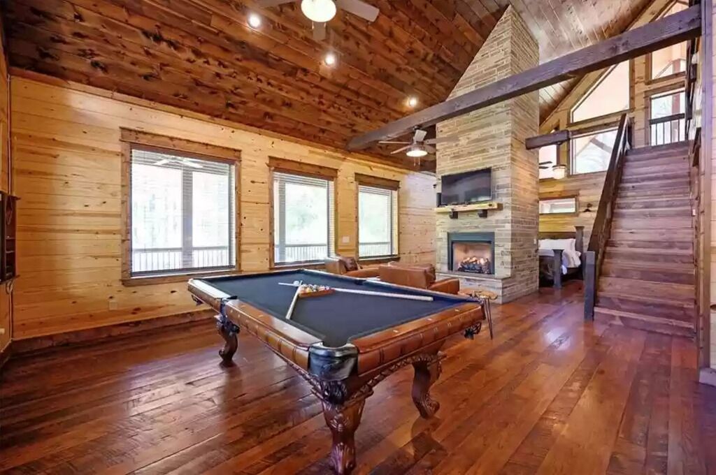 A wooden interior with a pool table, large windows, and a fireplace.