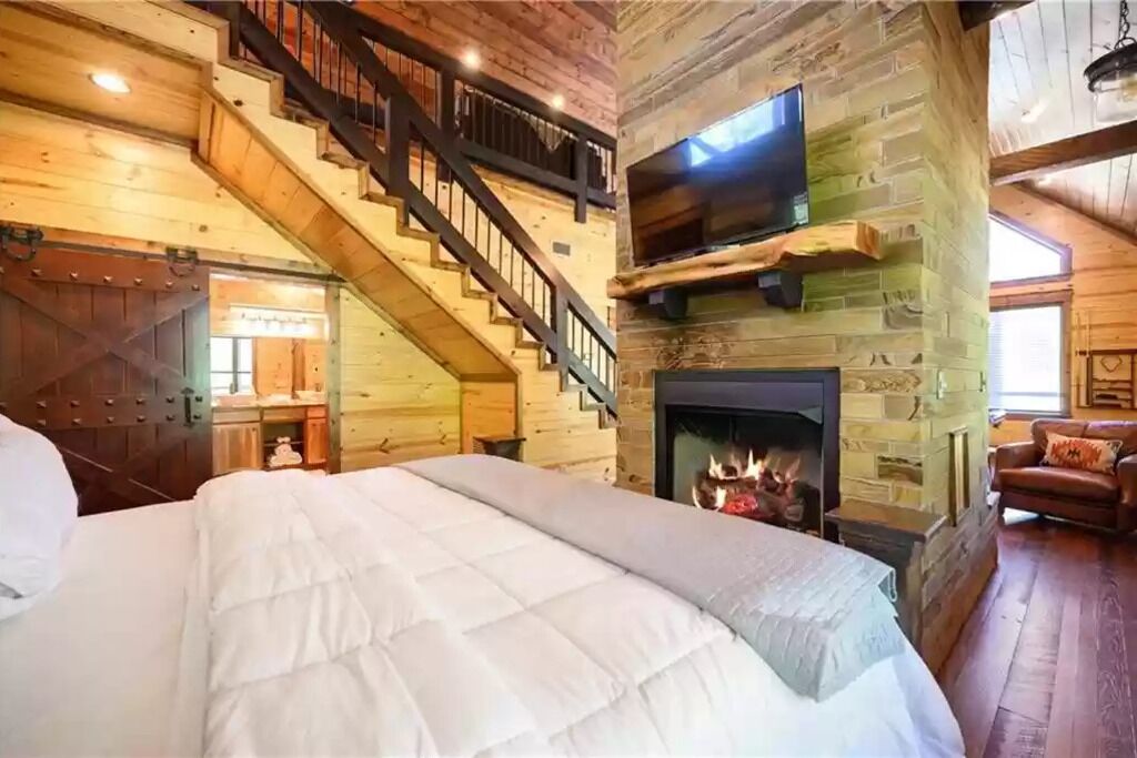 A log cabin-style bedroom with a large bed, a fireplace, a TV mounted on a stone wall, and a staircase leading to an upper level.