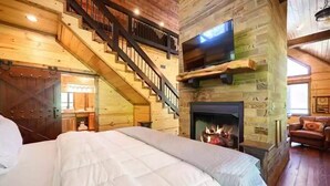 A log cabin-style bedroom with a large bed, a fireplace, a TV mounted on a stone wall, and a staircase leading to an upper level.