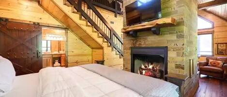 A log cabin-style bedroom with a large bed, a fireplace, a TV mounted on a stone wall, and a staircase leading to an upper level.
