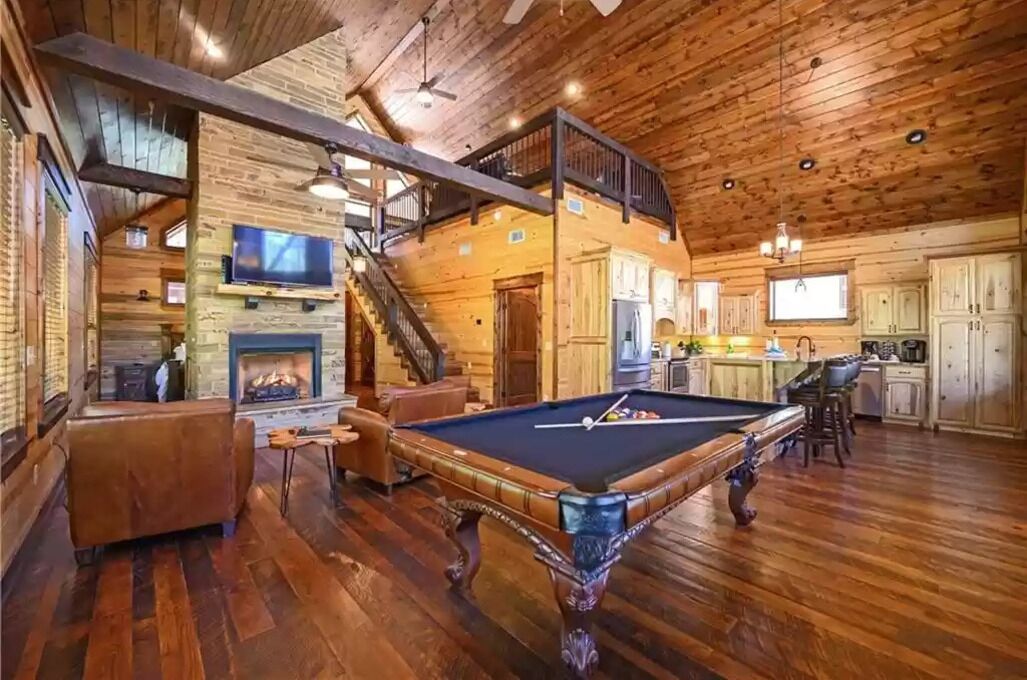A spacious wooden cabin with a pool table, a fireplace, and a kitchen area.