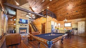 A spacious wooden cabin with a pool table, a fireplace, and a kitchen area.