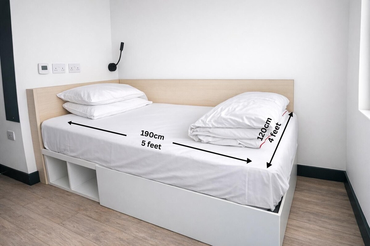 Habitación con una cama individual, cabecera de madera, ropa de cama blanca y un pequeño compartimento para guardar objetos debajo de la cama.