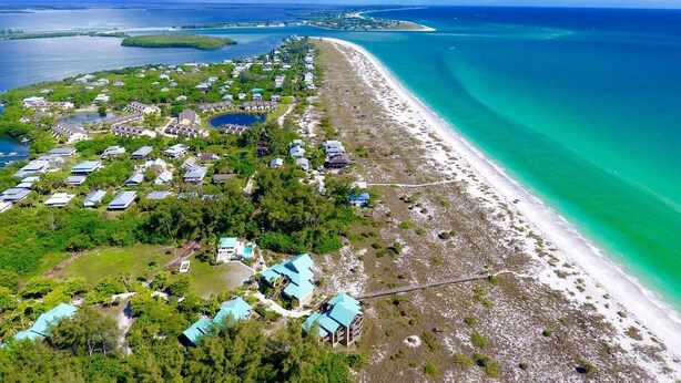 Vista aérea de un complejo turístico costero con propiedades frente a la playa, un puerto deportivo y aguas turquesas cristalinas.