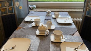 Une table de petit-déjeuner dressée avec des assiettes, des bols, des tasses et des couverts, donnant sur un champ verdoyant par la fenêtre.
