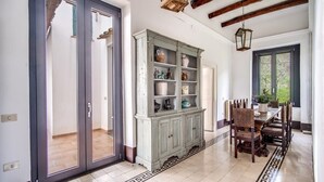 Uma ampla área de jantar com uma mesa grande e cadeiras, um armário com objetos decorativos, e vista para o exterior pela janela.