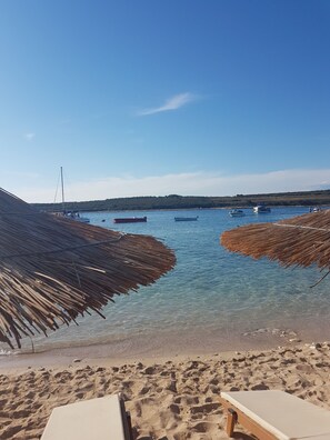 A beach with clear water, boats, and thatched umbrellas.