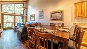 A dining area with a wooden table set for six, surrounded by chairs, with a view of trees through large windows. - Townhouse at the Woodlands 235 (Mammoth Lakes)