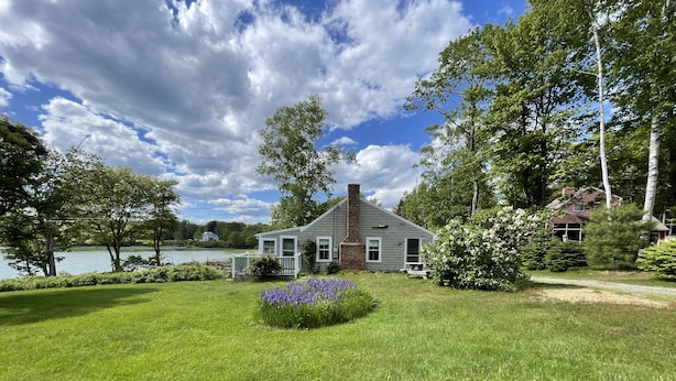 A house with a well-maintained lawn, a small garden with purple flowers, and a view of a lake. - Quaint Retro Ash Cove Cottage (Harpswell)