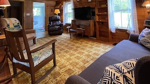 A cozy cabin interior with a wooden ceiling, a ceiling fan, a television, a fireplace, and a large window. - Quaint Retro Ash Cove Cottage (Harpswell)