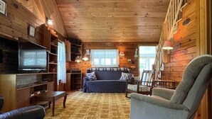A cozy cabin interior with a wooden ceiling, a ceiling fan, a television, a sofa, a chair, and a patterned rug. - Quaint Retro Ash Cove Cottage (Harpswell)