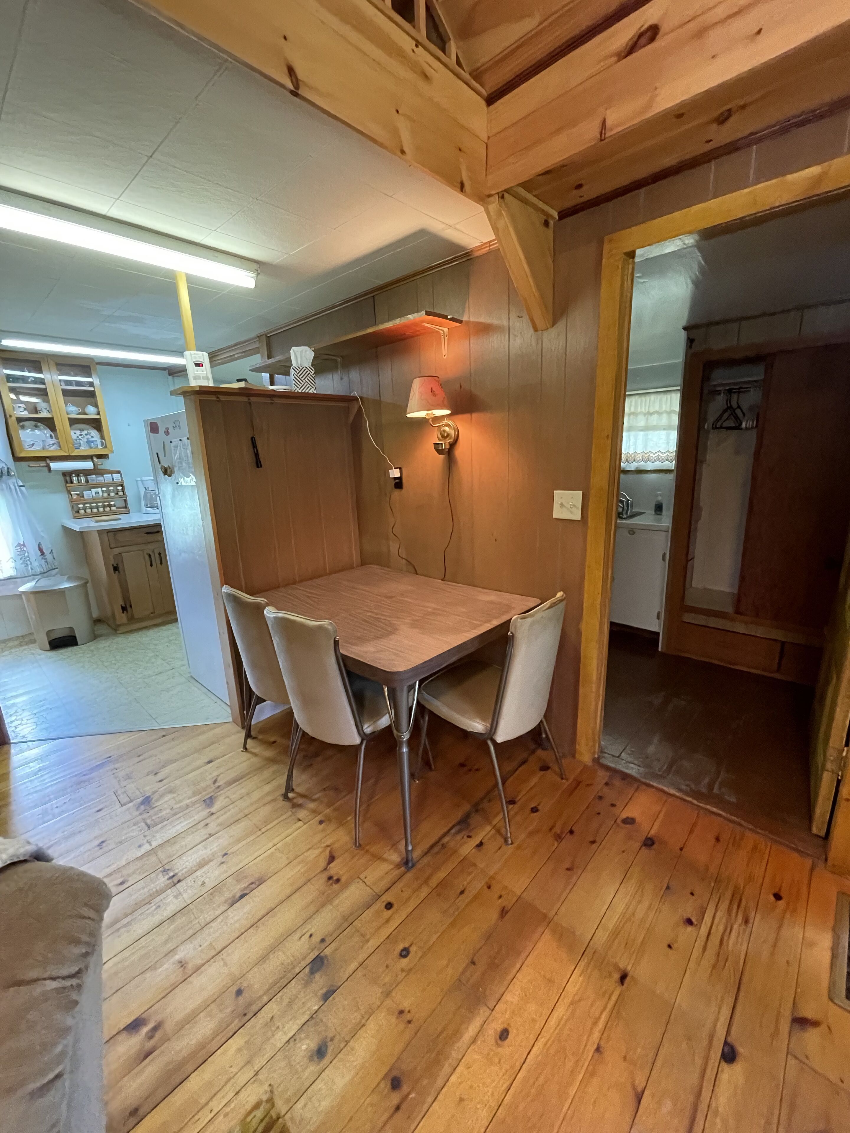 A wooden dining table with four chairs, a wall-mounted lamp, and a kitchen area visible in the background.