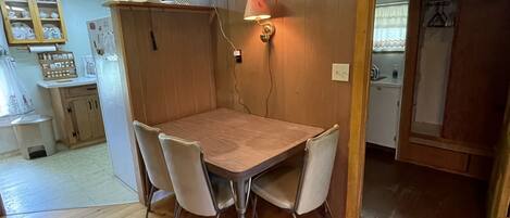 A wooden dining table with four chairs, a wall-mounted lamp, and a kitchen area visible in the background.
