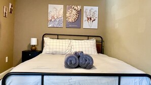 Cama bem arrumada com duas toalhas enroladas, mesa de cabeceira com abajur e três gravuras de plantas emolduradas na parede.