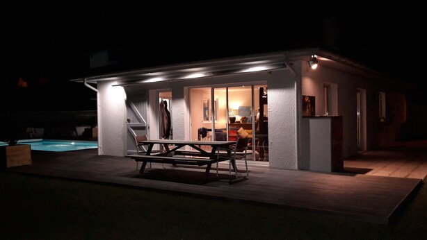 A small, well-lit outdoor patio area with a wooden table and chairs, a pool, and a house with a glass door leading to a living space.