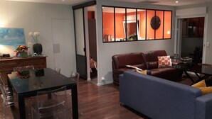 A living room with a brown leather sofa, a blue sofa, a dining table with chairs, and a TV stand with a flat-screen TV.