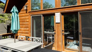 Una cabaña de madera con un espacio de terraza que incluye una mesa, sillas, un paraguas verde y un cartel con las normas de la terraza.