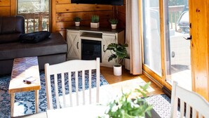 Interior acogedor de una cabaña con techo de madera, televisor de pantalla plana en la pared, sofá, mesa de comedor y ventana con vista al exterior.