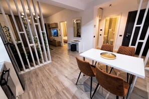 A modern dining area with a white table and brown chairs, a kitchen in the background, and a decorative plant barrier.