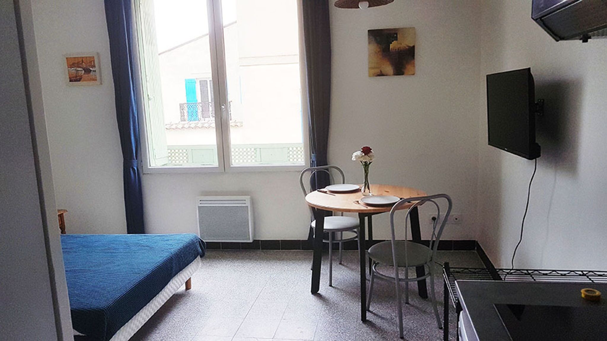A small, well-lit room with a single bed, a table with chairs, a TV mounted on the wall, and a window with a view of a building outside.