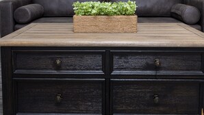 A dark brown leather sofa with a woven basket containing green plants on a wooden coffee table. Below the sofa is a dark wooden dresser with two drawers.