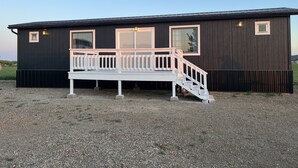 A small, dark wooden cabin with a white deck and a staircase leading to a windowed porch. - Sleeps 8 — Bunkhouse (Platte)