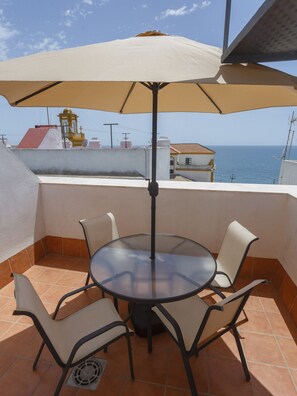 A round table with four chairs and a large umbrella on a tiled patio overlooking the sea.