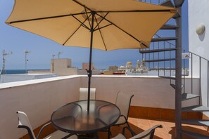 A rooftop terrace with a table and chairs, an umbrella, and a view of the sea and buildings in the distance.