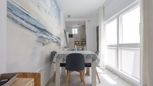 Uma sala de jantar com mesa posta para quatro pessoas, um quadro na parede e vista para a cozinha ao fundo.