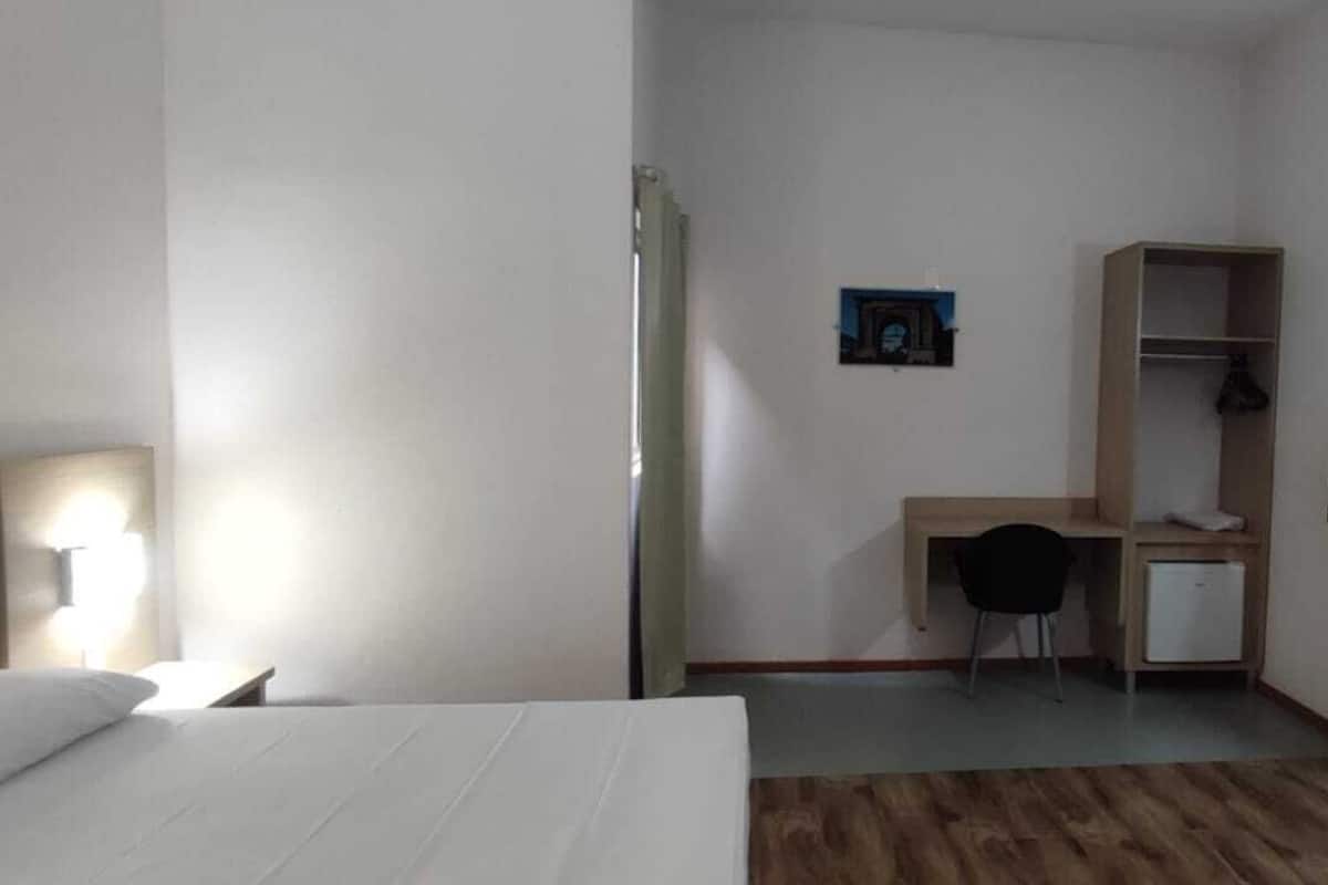 A hotel room with a bed, a desk, a chair, a small refrigerator, a wardrobe, a mirror, and a wall-mounted TV.
