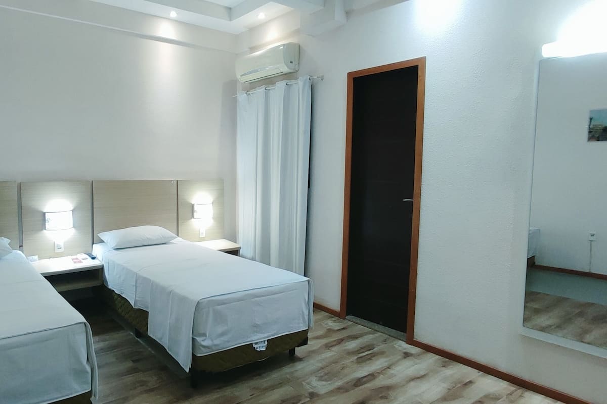 A hotel room with two single beds, a nightstand with a lamp, an air conditioning unit, a door with a mirror, and a small picture on the wall.
