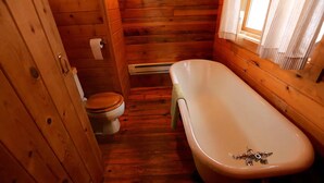 A wooden cabin bathroom with a toilet, a bathtub, and a window with curtains.