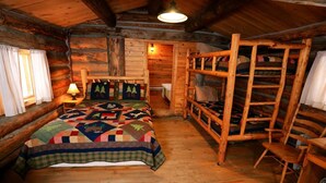 A rustic cabin bedroom with a wooden bed, a quilt, a wooden ladder, and a small table with a lamp.
