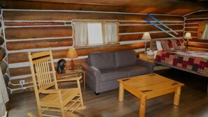 L’intérieur d’une cabane en rondins, avec un fauteuil à bascule en bois, un canapé gris, une table basse en bois et une courtepointe.