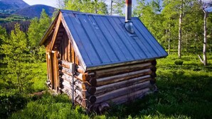 A rustic log cabin with a metal roof and chimney, surrounded by lush greenery and mountains in the background. - Porcupine Cabin - A Classy-Rustic Gem (Clark)