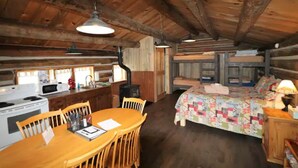 A rustic cabin interior with a wooden dining table, chairs, a kitchen with a stove and microwave, a bed with a quilt, and a wooden ceiling. - Porcupine Cabin - A Classy-Rustic Gem (Clark)