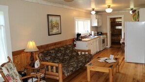 A cozy living area with a wooden sofa, a small table, a lamp, and a kitchen in the background. - Caron House - A Well-Equipped Square Log Cabin! (Clark)