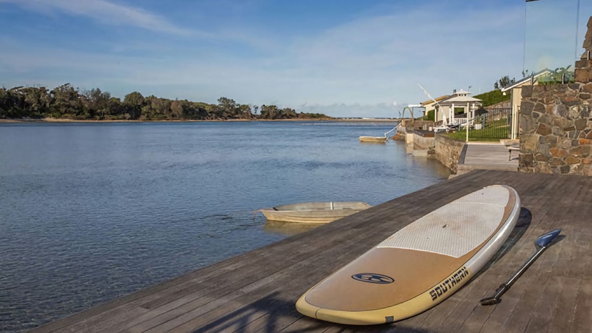 Une jetée en bois, avec une planche de surf et une pagaie, donnant sur une rivière paisible, bordée d’un mur de pierres et avec un bâtiment en arrière-plan.